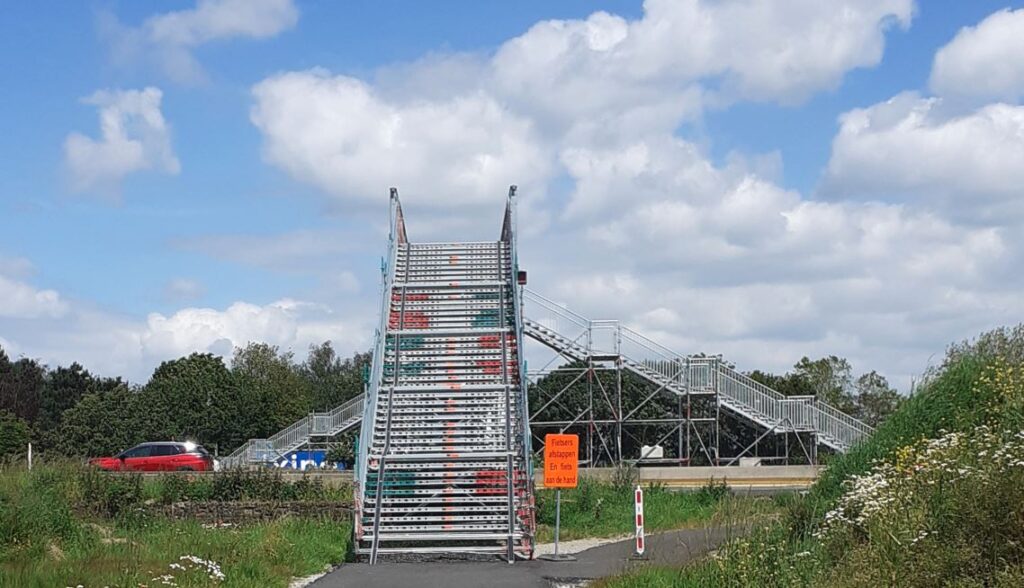 A very high and steep flight of stairs made of metal, intended for temporary use. It helps pedestrians crossing a highway. there is a small an orange sign tells: bicyclists, descend and take your bike by the hand